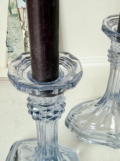 Clear cut-glass candlestick holders with a dark brown candle in the foreground and another holder partially visible on the right, set against a light neutral background.