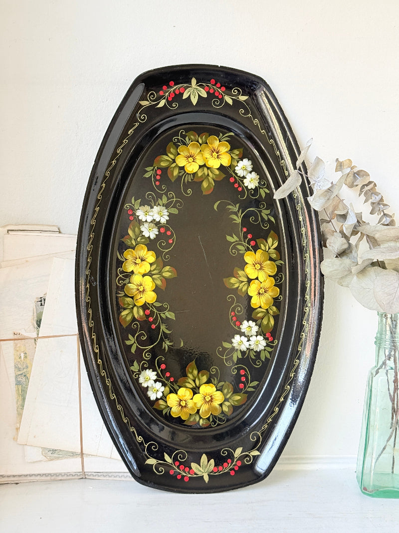 An oval black decorative tray with hand-painted yellow flowers, green leaves, and small red berries, styled on a white surface next to a glass vase with dried flowers.