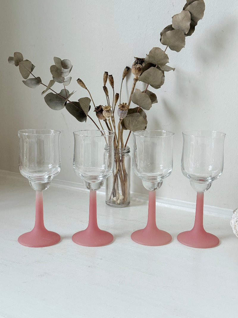 Set of four small vintage wine glasses with clear bowls and pink stems photographed on a white surface.