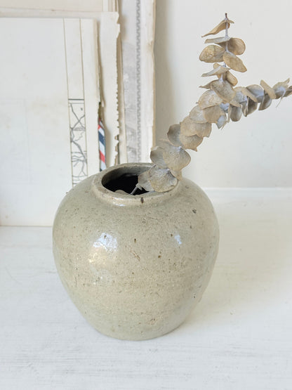 A small round ceramic vase with a speckled light beige-gray glaze, holding a dried eucalyptus branch, set against a soft white background with vintage-style papers.