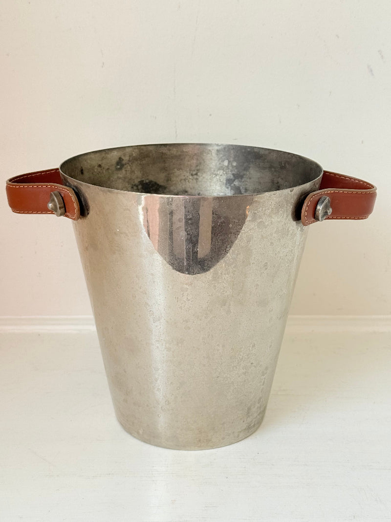 Vintage metal wine cooler with leather handles and tapered form, photographed on white surface.