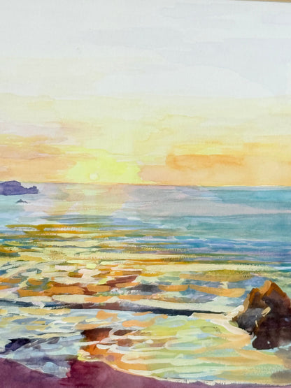 Framed coastal watercolour of sunset over sea with rocks in foreground, in gilt frame with pale mount, photographed on white surface.