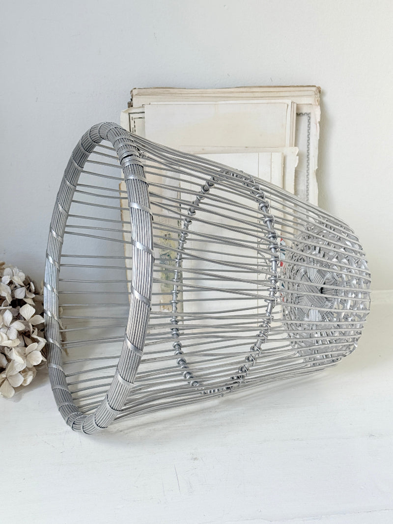 A silver metal wire basket-style wall decor piece with circular wire patterns, displayed against a white wall with a vintage-style frame behind it.
