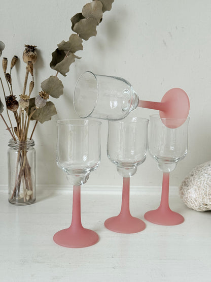 Set of four small vintage wine glasses with clear bowls and pink stems photographed on a white surface.