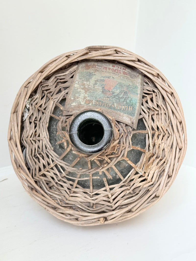 antique French demi john olive oil jar in wicker basket with green glass bottle and painted metal label, displayed on white surface