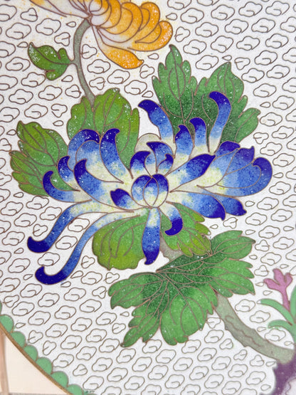 Close-up of a decorative floral artwork showing a blue and white chrysanthemum flower with green leaves on a white patterned background.