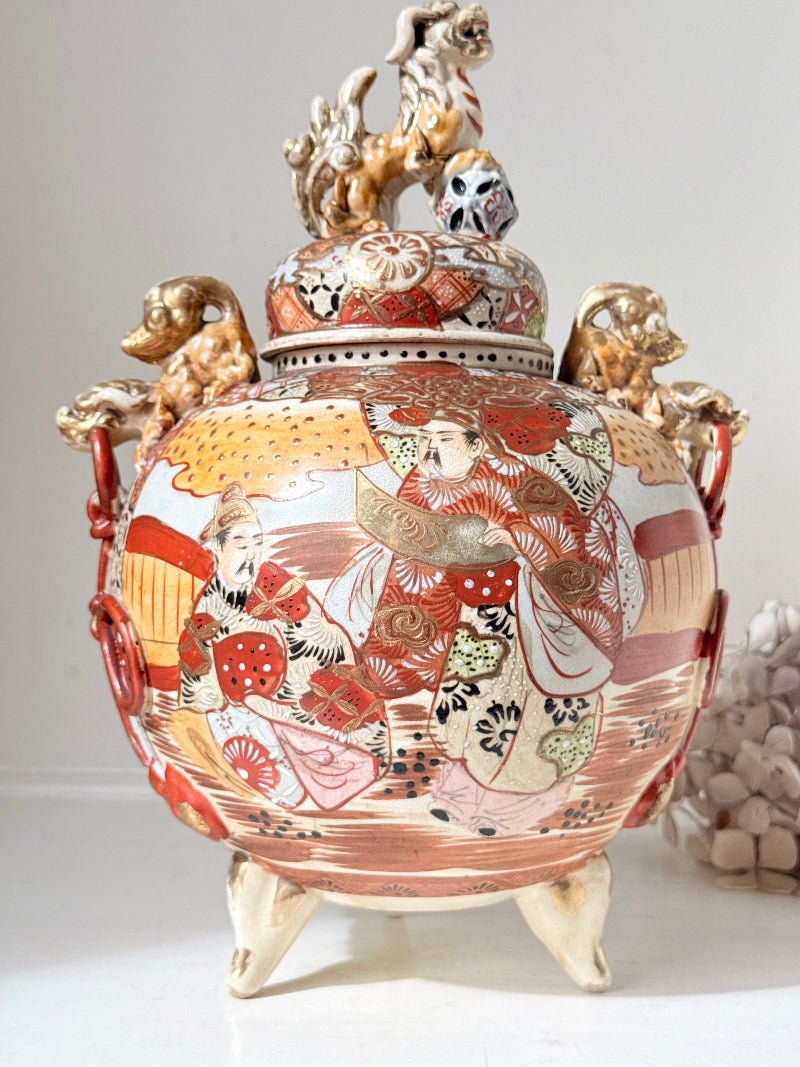 An ornate Japanese-style ceramic lidded vase with a hand-painted kimono and figure design in red, gold, and cream, featuring three gold-colored dog figures on the sides and a dragon figure on top of the lid.