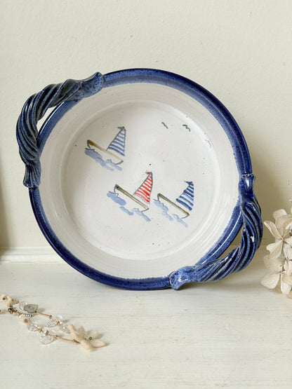 A round white ceramic bowl with a dark blue glazed rim and twisted side handles, decorated with three small sailboats on the inside. The bowl is styled on a white surface with dried flowers and a pearl strand against a cream background.