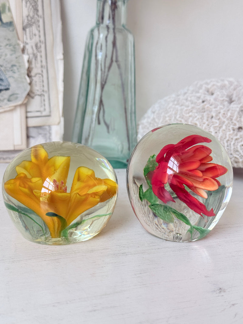 Pair of orange and yellow flower glass paperweights with green leaves, photographed on a white surface