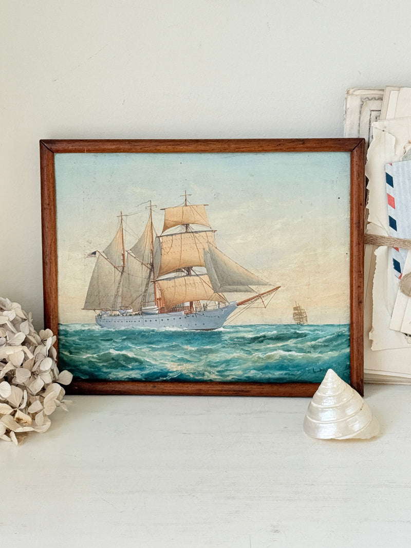 A framed vintage-style painting of a sailing ship on blue ocean waves, displayed on a white surface with decorative seashells and framed art around it.