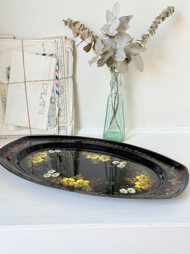 A black oval decorative tray with yellow and white floral patterns, styled on a white surface next to a vintage envelope bundle and a glass vase with dried leaves.