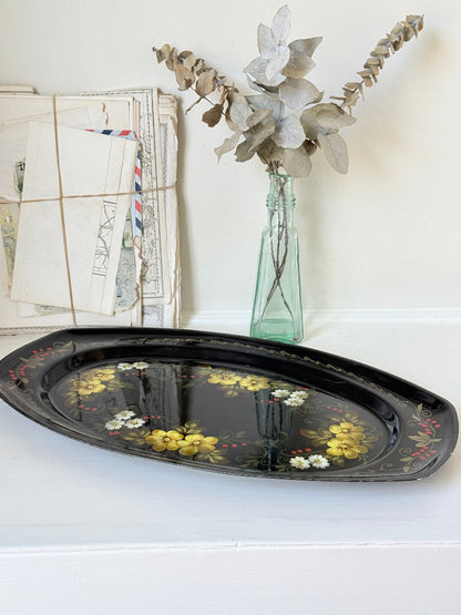 A black oval decorative tray with yellow and white floral patterns, styled on a white surface next to a vintage envelope bundle and a glass vase with dried leaves.