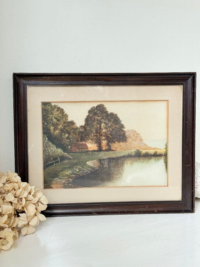 Antique framed rural watercolour of river, trees, and red-roofed building, in dark wooden frame with cream mount, on white surface.