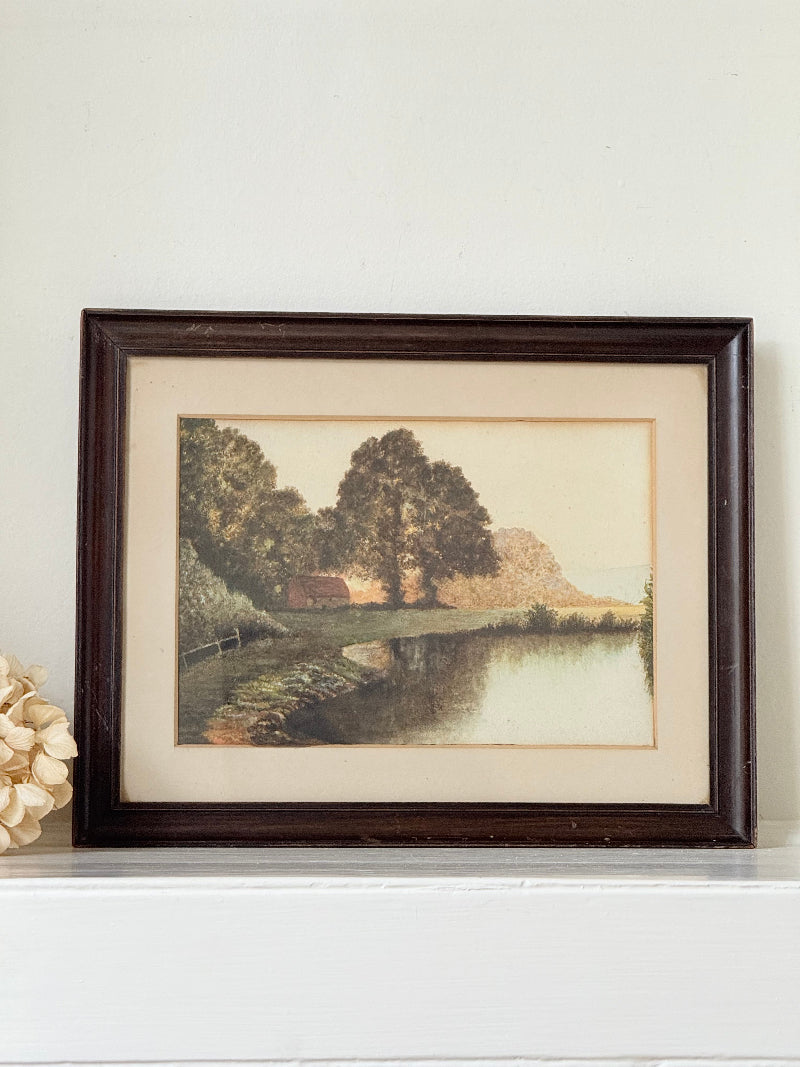 Antique framed rural watercolour of river, trees, and red-roofed building, in dark wooden frame with cream mount, on white surface.