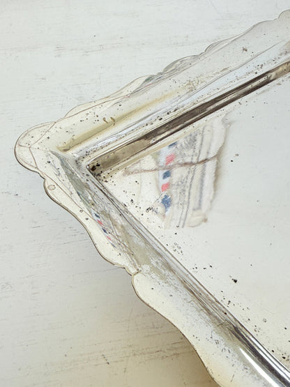 A vintage silver-toned metal tray with a scalloped edge, showing signs of age, wear, and rust, resting on a distressed white painted wooden surface.
