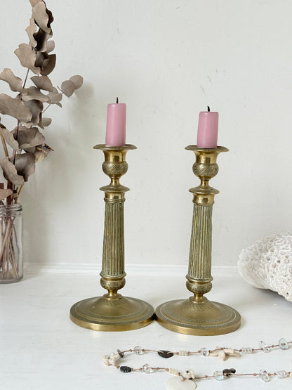Pair of antique brass Empire style candlesticks photographed on a white surface with fluted columns and circular bases holding pink taper candles