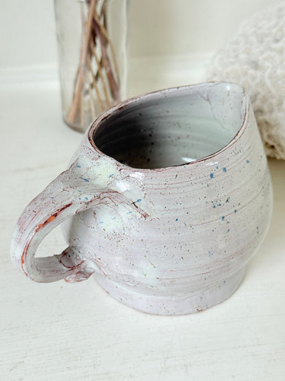 Studio pottery milk jug with soft pink and grey speckled glaze, photographed on a white surface