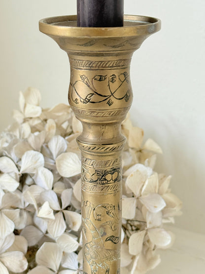 Pair of tall Indian engraved brass candlesticks in the Benares style with floral detailing and black taper candles on a white surface