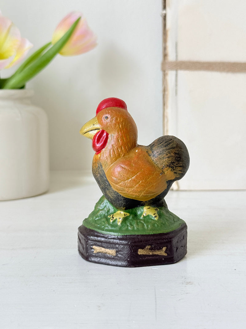 A vintage hand-painted cast iron hen figurine on a dark base, displayed on a white surface with a blurred white vase of tulips in the background.