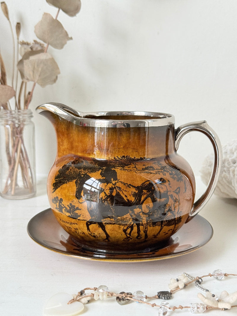 Antique Ridgways stoneware creamer jug and saucer photographed on a white surface in amber treacle glaze with black coaching scenes and silver lustre rim