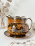 Antique Ridgways stoneware creamer jug and saucer photographed on a white surface in amber treacle glaze with black coaching scenes and silver lustre rim