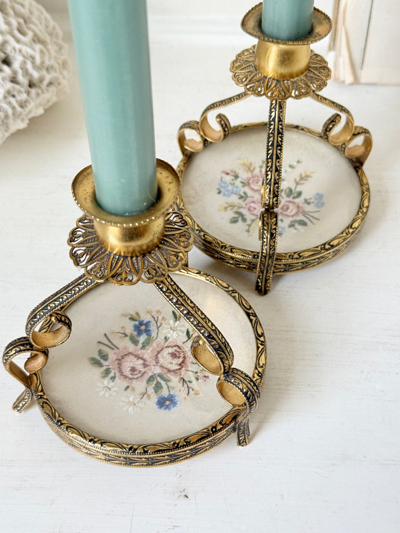 Two ornate gold candlestick holders with floral porcelain bases, holding light blue candles on a white wooden surface.
