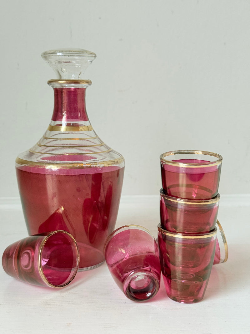 Vintage red printed glass cocktail set with clear glass decanter and six small glasses with gilt band rims, photographed on a white surface