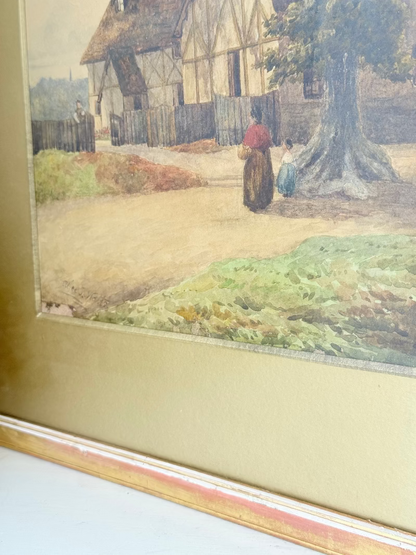 Antique framed watercolour of English village street with thatched cottages and figures, in gilt frame with wide mount, on white surface.