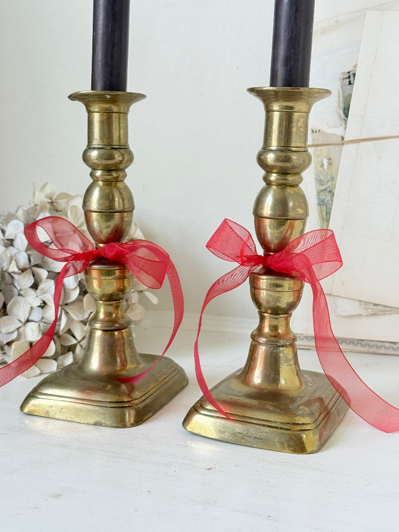 Pair of turned brass candlesticks with square stepped bases and black taper candles on a white surface, styled with dried flowers