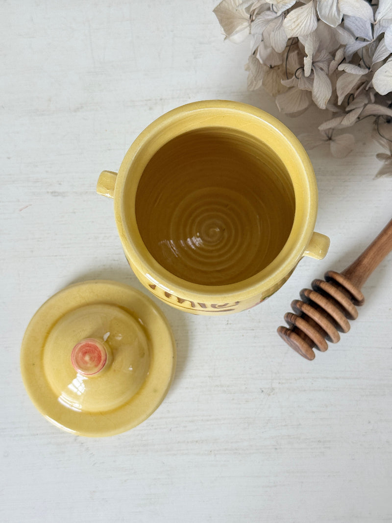 A yellow ceramic honey pot with its lid off, showing a honey-colored liquid inside, styled on a white wooden surface with a wooden honey dipper and dried flowers.