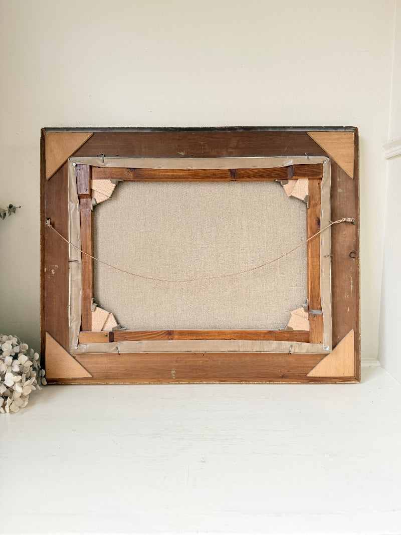 The back view of a large wooden picture frame with a linen-wrapped canvas backing, resting on a white surface against a light beige wall.