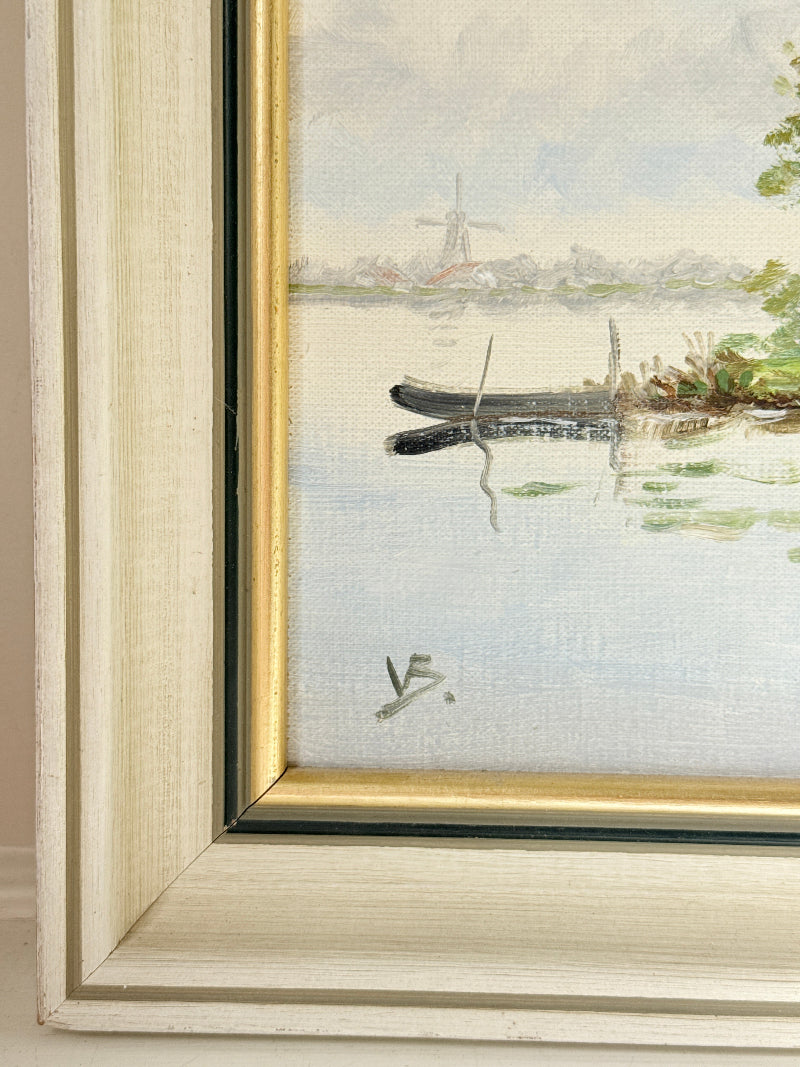 Close-up of a framed landscape painting showing a painted water scene with a small boat, distant windmill, and pale sky, bordered by a light wood frame with a gold inner trim.