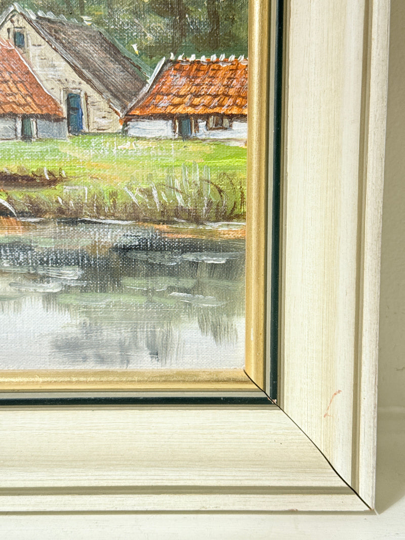 Close-up of a framed painting showing a rural landscape with two cottages, orange-tiled roofs, green grass, and a reflective pond. The frame is a light cream wood with a gold inner border.