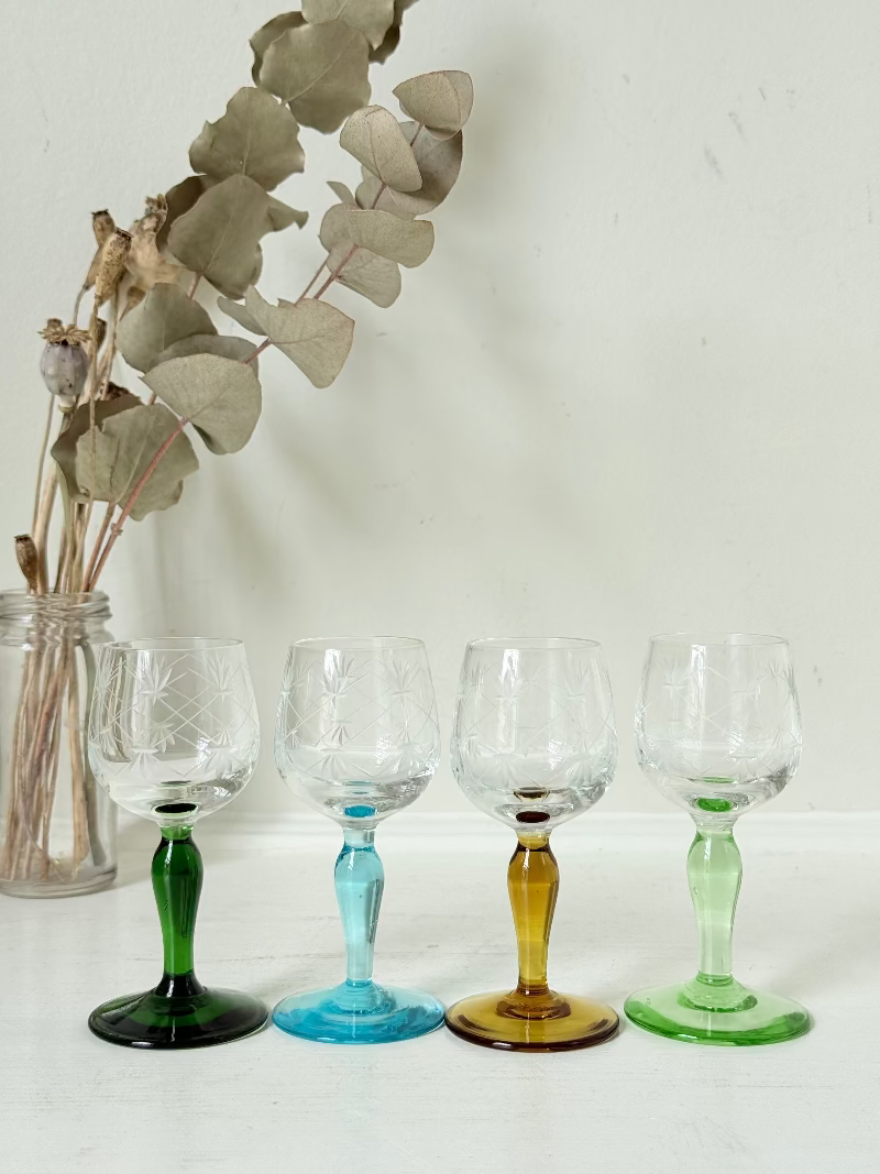 Vintage set of four etched liqueur glasses with colourful stems in green, amber, blue, and turquoise, photographed on a white surface