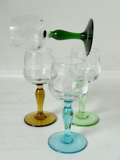 Vintage set of four etched liqueur glasses with colourful stems in green, amber, blue, and turquoise, photographed on a white surface