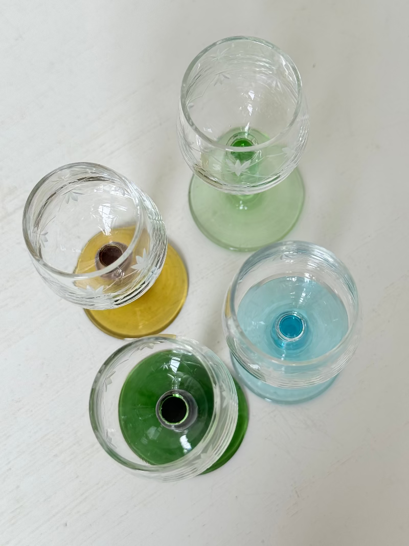 Vintage set of four etched liqueur glasses with colourful stems in green, amber, blue, and turquoise, photographed on a white surface