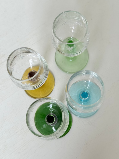 Vintage set of four etched liqueur glasses with colourful stems in green, amber, blue, and turquoise, photographed on a white surface