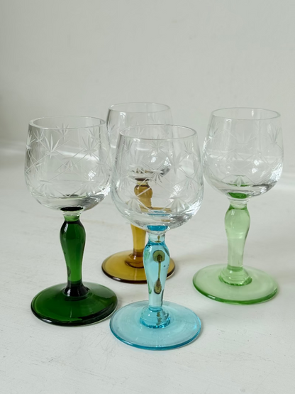 Vintage set of four etched liqueur glasses with colourful stems in green, amber, blue, and turquoise, photographed on a white surface
