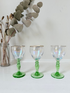 Vintage trio of iridescent shot glasses with green stems and gilt rims, photographed on a white surface