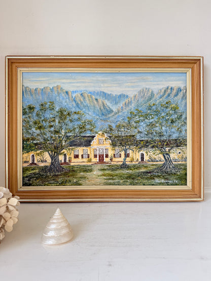 A framed landscape painting of a yellow building set among green trees with blue-gray mountains in the background, displayed on a white surface with a decorative shell and dried flowers nearby.