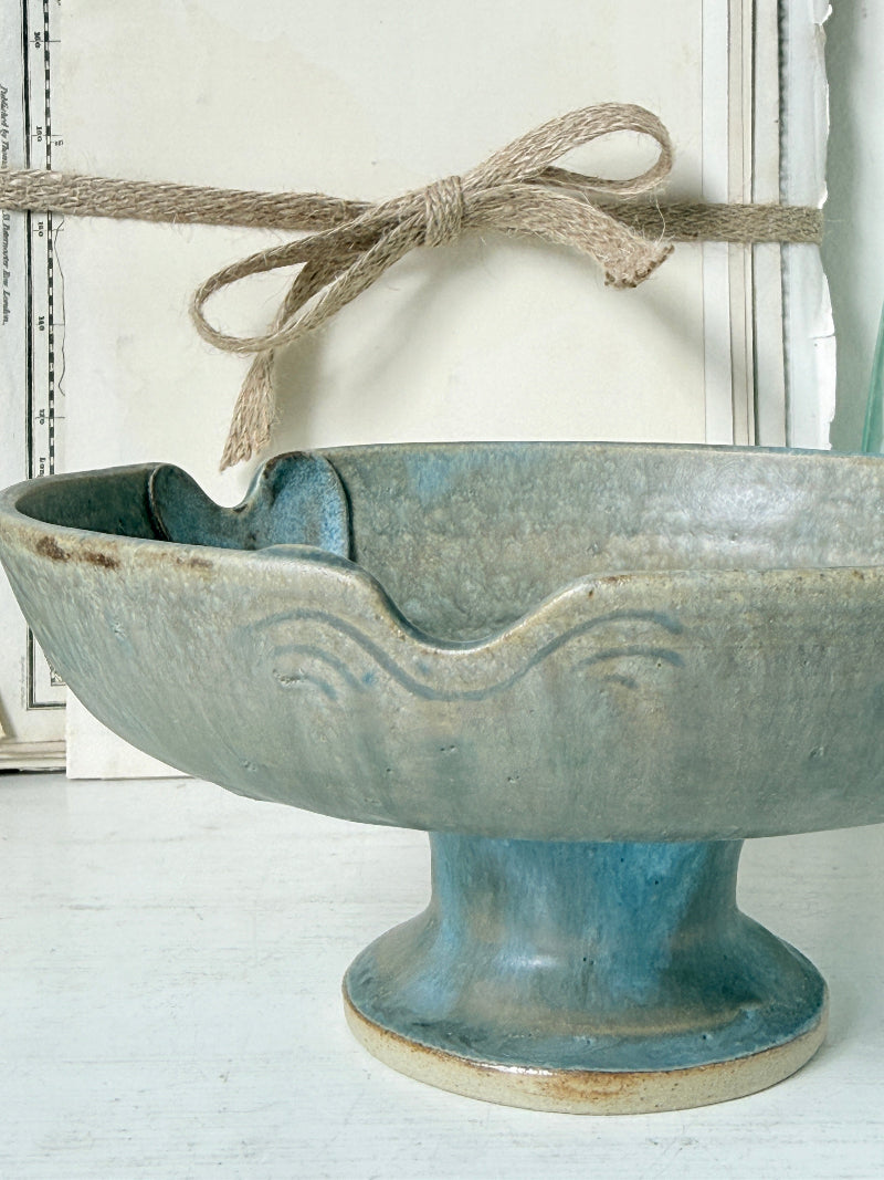 A rustic ceramic pedestal bowl with a flared rim and embossed wave pattern, featuring a blue-green glazed finish with natural beige edges. A piece of jute twine is tied around the rim in the background against a white paneled wall.