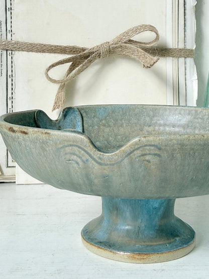 A rustic ceramic pedestal bowl with a flared rim and embossed wave pattern, featuring a blue-green glazed finish with natural beige edges. A piece of jute twine is tied around the rim in the background against a white paneled wall.