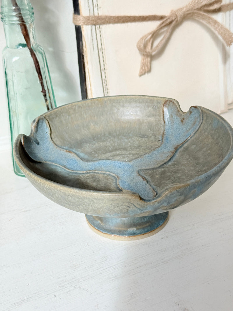 A handmade ceramic pedestal bowl with a rustic, hand-painted finish in muted blue-gray and beige tones, styled on a white wooden surface with a glass bottle and decorative background elements.