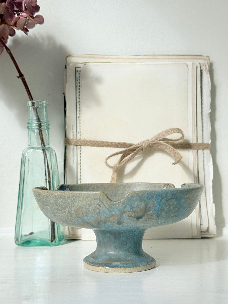 A blue-gray ceramic pedestal bowl with a distressed finish, styled in a rustic setting with a vintage book wrapped in twine and a small glass bottle.