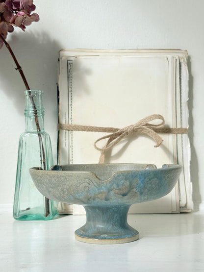 A blue-gray ceramic pedestal bowl with a distressed finish, styled in a rustic setting with a vintage book wrapped in twine and a small glass bottle.