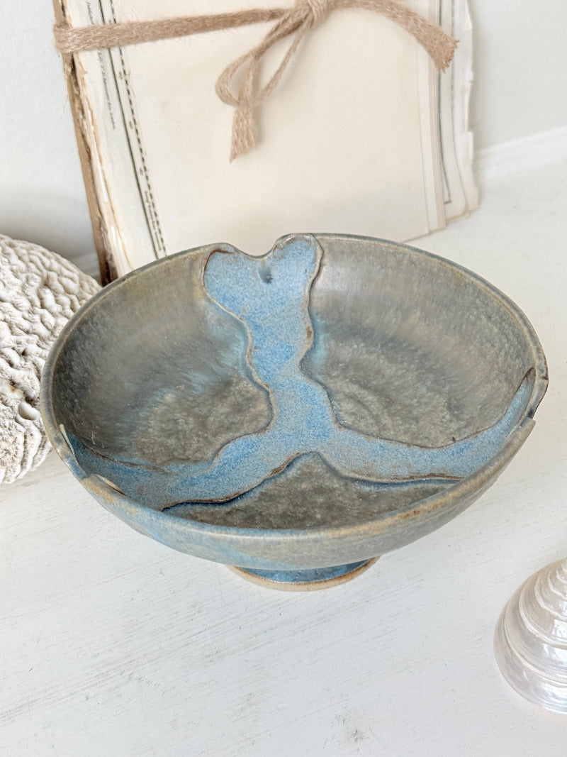 A handmade ceramic bowl with a rustic, hand-painted blue and gray glaze, styled on a white wooden surface with decorative background elements.