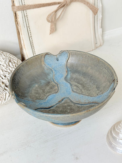 A handmade ceramic bowl with a rustic, hand-painted blue and gray glaze, styled on a white wooden surface with decorative background elements.