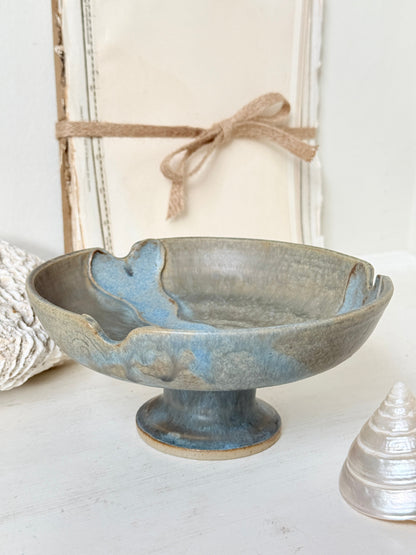 A handmade ceramic pedestal bowl with a distressed blue and gray glaze, styled on a white surface with decorative props including a wooden frame, burlap ribbon, and seashells.