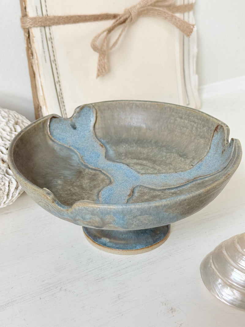 A rustic ceramic pedestal bowl with a blue and gray glaze, styled on a white surface with a linen box and jute ribbon in the background.