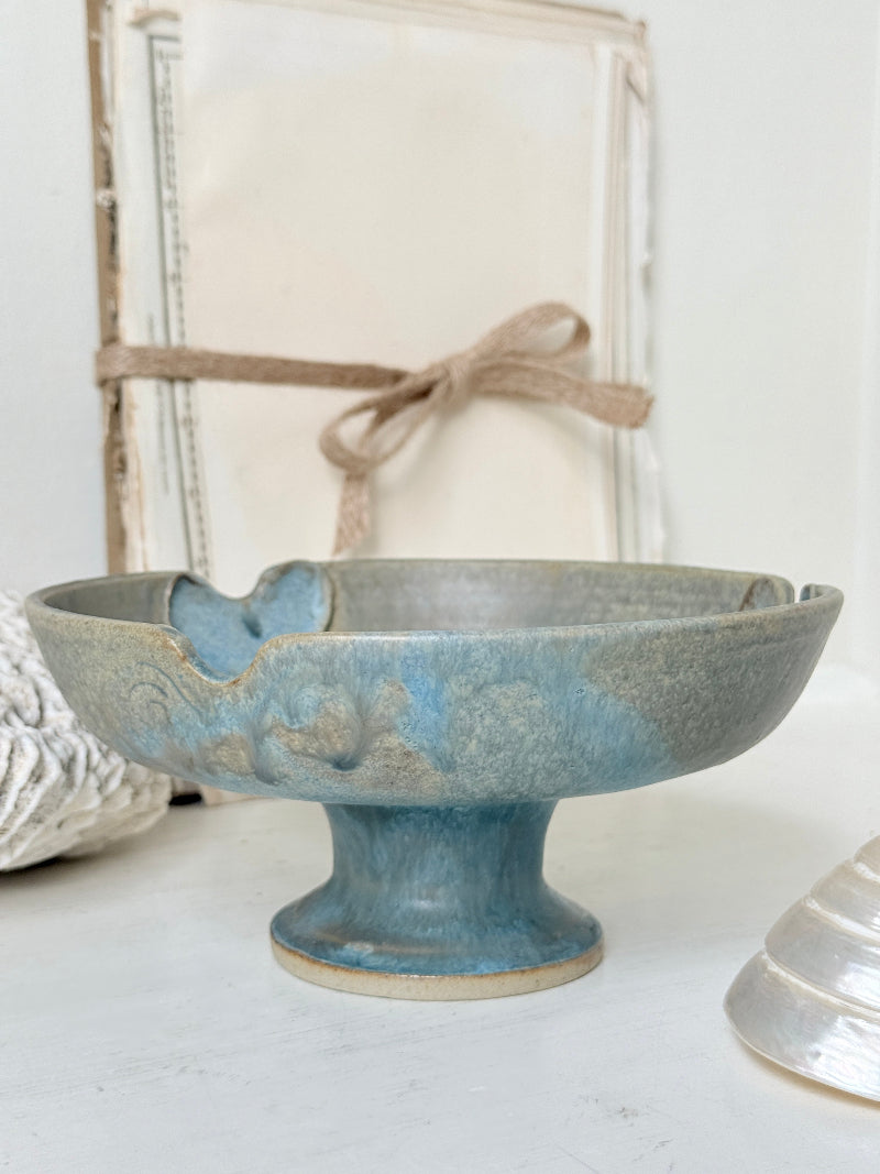 A handmade ceramic pedestal bowl with a distressed blue and beige glaze, styled on a white surface with a rustic, vintage-themed background.